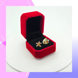 A pair of small starfish & fish asymmetrical stud earrings displayed in an open red velvet ring box on a white surface against a white background.