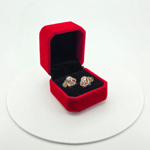 A pair of colorful clownfish stud earrings displayed in an open red velvet ring box on a white circular platform against a white background.